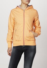 Giacca leggera con cappuccio in arancione pastello con zip frontale, polsini elasticizzati e tasche laterali. Tessuto texture e vita arricciata.