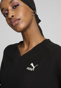 Črn pleteni top z V-izrezom, ki ima teksturiran vzorec in bel logotip Puma. Zaključen z rebrastimi detajli na izrezu in rokavih.
