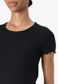 Musta ribaatattu paita, jossa on pyöreä pääntie ja lyhyet hihat, joiden reunassa on aallotettu viimeistely. Kangas vaikuttaa pehmeältä ja hieman teksturoidulta.