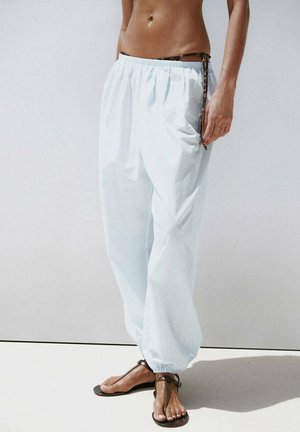 Persona que lleva pantalones blancos holgados con tobillos elásticos y sandalias marrones, de pie frente a un fondo liso y claro.