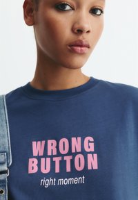 Blaues Baumwoll-T-Shirt mit kurzen Ärmeln, das "WRONG BUTTON" in fettgedruckten pinken Buchstaben und "right moment" in kleinerer Schrift darunter zeigt.