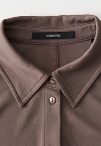 Polo shirt marron avec un col pointu, doté d'une patte de boutonnage et d'une texture subtile. L'étiquette indique "un jour". Les boutons sont pâles, assortis au tissu.