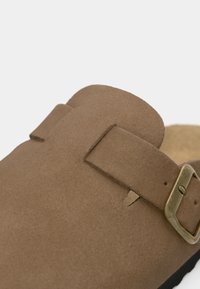 Puha barna velúr slip-on cipő állítható pánttal és sárgaréz csattal, lekerekített orral és párnázott talppal.