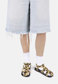 Sandalias de estampado de leopardo con suelas negras, acentos en dorado y negro, llevadas con calcetines blancos hasta el tobillo. Pantalones cortos de mezclilla desgastados de color azul claro.