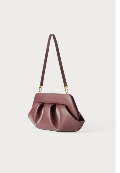 Μικρή μωβ δερμάτινη τσάντα ώμου με μαζεμένες πτυχές και λεπτό λουρί που είναι συνδεδεμένο με χρυσό υλικό, εκτεθειμένη σε απλό φόντο.