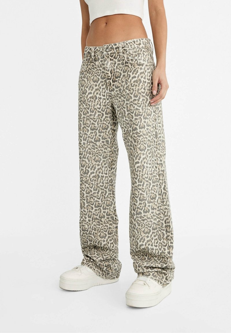 Femme portant un pantalon ample taille haute à imprimé léopard, des baskets blanches à plateformes et un top blanc court, debout sur un fond blanc.