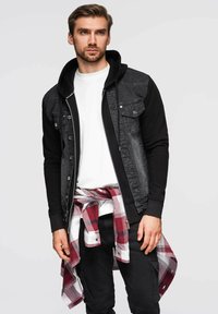 Giacca in denim nera con cappuccio, pannelli grigi sulle spalle, due tasche sul petto, indossata sopra una camicia bianca, pantaloni neri e una camicia a quadri rossa legata in vita.