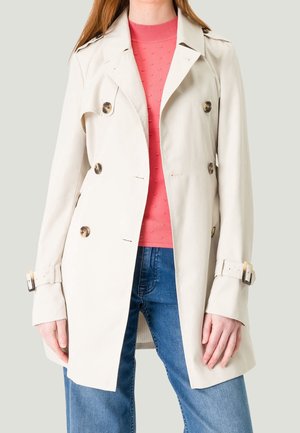 Femme portant un trench-coat beige à double boutonnage sur un pull texturé corail et un jean bleu, debout devant un fond uni.
