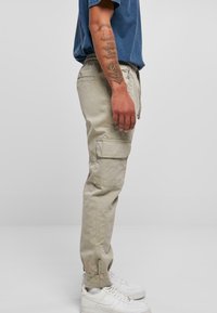 Beige cargo bukser med en tapered design, elastisk talje, sidelommer og manchetter ved anklerne, parret med hvide sneakers og en blå skjorte.