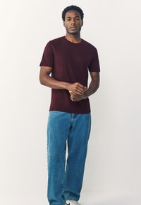 Tumepunane lühikeste varrukatega t-särk, kombineeritud helesiniste teksapükstega. Kangas tundub pehme, lõige on mugav ja vaja ei ole nähtavaid mustreid ega dekoratsioone.