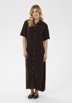 Žena se světlými vlasy v černých maxi šatech s krátkými rukávy a černých pletených balerínkách, stojící před bílým pozadím.