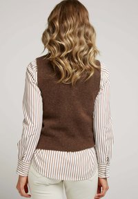 Gilet en laine marron au design ajusté, porté sur une chemise rayée blanche et marron, associé à un pantalon de couleur claire. Tissu en tricot texturé.