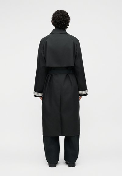 Personne aux cheveux bouclés et foncés portant un long trench-coat noir avec des détails à carreaux sur les poignets, debout de dos sur un fond blanc.