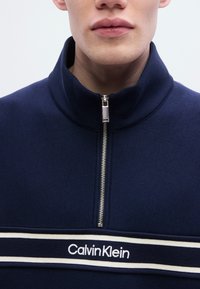 Φούτερ με φερμουάρ σε ναυτικό μπλε, με ψηλό γιακά, λευκή και ναυτική ριγέ ταινία με το λογότυπο "CalvinKlein" και ομαλή υφή υφάσματος.
