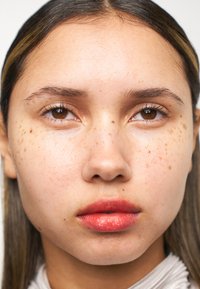 Närbild på ett ansikte med ljusbrun hud, fräknar, rakt svart hår och en mjuk korallfärgad läppfärg. Ögonen är mörka och mandelformade.