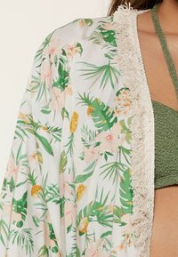 Kimono floral con patrones tropicales en rosa y verde, tejido semi-translúcido, con bordes de flecos en blanco y un frente abierto.