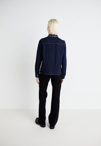 Denim jacket in dark blue with a solid collar featuring silver studs, long sleeves with button cuffs, paired with black pants.