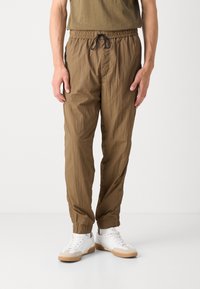 Leichte braune Hose mit elastischem Bund und Kordelzug, mit schmal zulaufendem Bein und glatter Textur. Getragen mit weißen Sportschuhen.