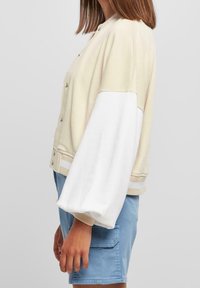 Světle béžová bomber bunda s bílými blokovými rukávy, žebrovanými lemy a zapínáním na patentky; volný střih a měkká textura.