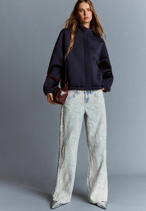 Navy oversized sweatshirt with high collar, paired with light-wash wide-leg jeans and silver pointed-toe heels. Small burgundy bag accent.