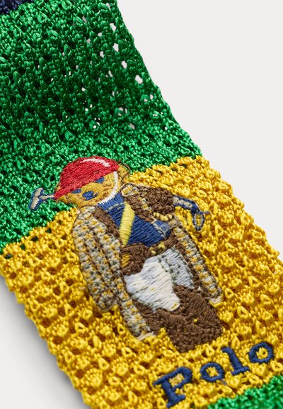 Pletená látka so zelenými a žltými časťami s vyšívaným medveďom, ktorý má na sebe červenú čiapku, pruhovanú bundu a modrú košeľu, s textom „Polo“ pod tým.