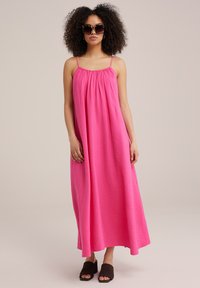 Rosa maxiklänning med justerbara spaghettistövlar, texturerad tyg, lös passform och flödande silhuett. Modell iklädd bruna sandaler och solglasögon.