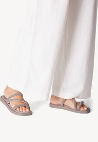 Sandali marroni chiari con due cinturini incrociati realizzati in materiale testurizzato, posizionati su una base piatta, abbinati a pantaloni bianchi a gamba larga.
