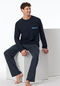 Camisa de manga larga azul marino con un bolsillo en el pecho de color azul claro, combinada con pantalones de pijama oscuros de cuadros con un patrón de cuadrícula. Modelo sentado sobre un cubo blanco.
