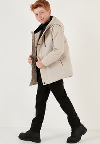 Beige vadderad jacka med huva, dragkedja fram, svarta detaljer och sidofickor, bärs över en svart topp och ihop med svarta byxor och stövlar.