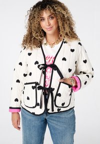 Vrouw met krullend haar, gekleed in een crèmekleurige jas met zwarte harten, roze manchetten, een witte graphic tee, blauwe spijkerbroeken en gouden sieraden, die zachtjes glimlacht.