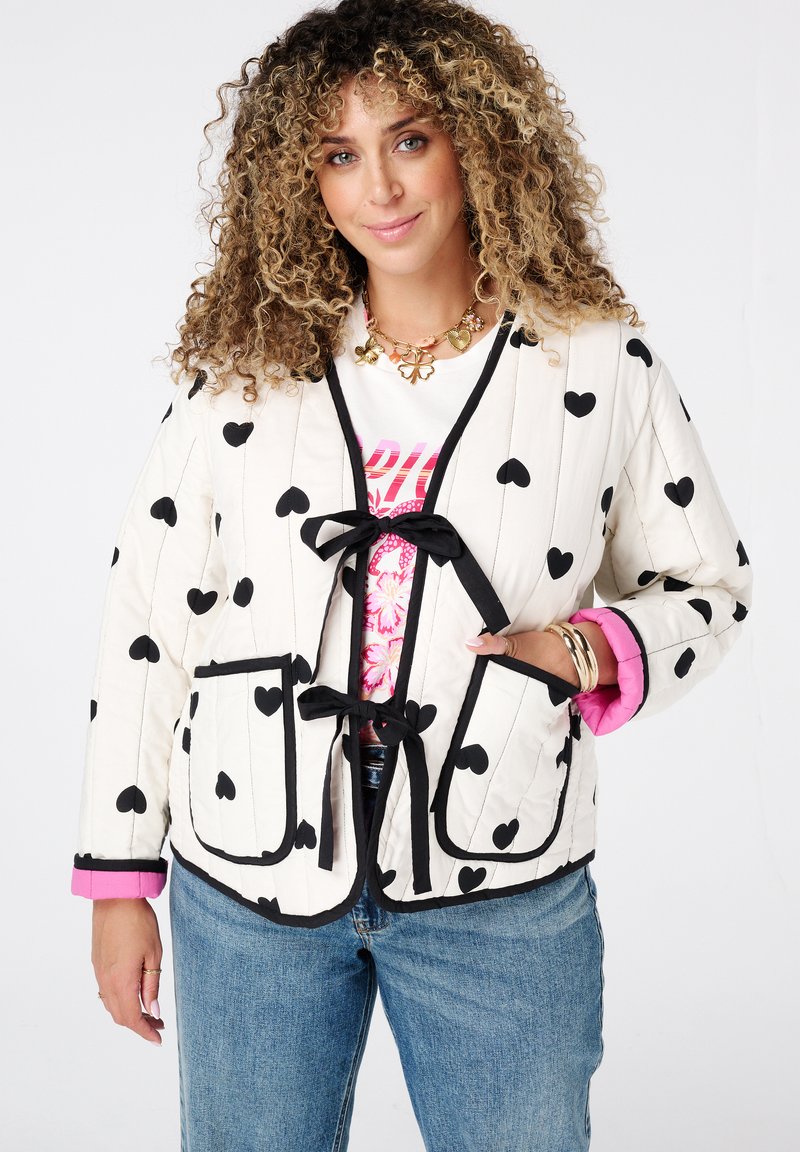 Vrouw met krullend haar, gekleed in een crèmekleurige jas met zwarte harten, roze manchetten, een witte graphic tee, blauwe spijkerbroeken en gouden sieraden, die zachtjes glimlacht.