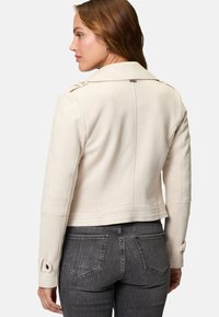 Giacca cropped beige chiara con una texture liscia e spalle strutturate. Presenta grandi bottoni e dettagli di cucitura sottili.