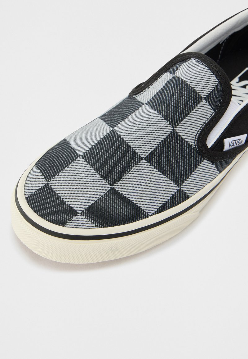 Zapatilla tipo slip-on en blanco y negro con un diseño de cuadros, parte superior de lona, acentos elásticos en los laterales y suela de goma blanca con ribete negro.