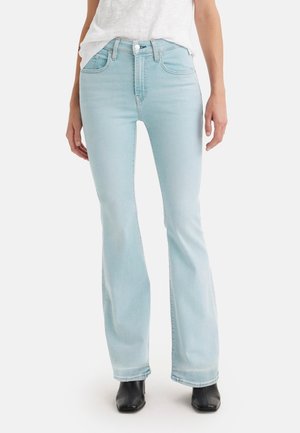 Kobieta w jasnoniebieskich dzwonowatych dżinsach z wysokim stanem, czarnych botkach na kwadratowym nosku oraz białej bluzce z krótkim rękawem, stojąca na białym tle.