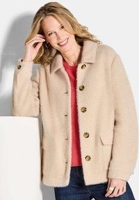 Beige wolblend jas met een geprofileerde kraag, knopenfront, twee zakken en textuurstof. Draagt over een rode trui en donkere spijkerbroek.