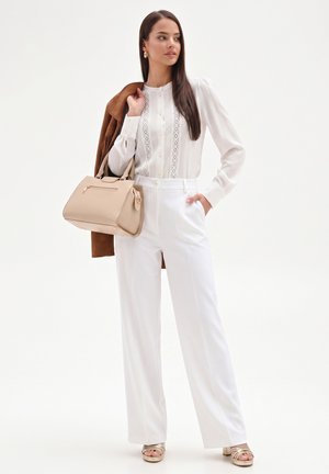 Donna che indossa una camicetta bianca e pantaloni, tiene una borsa beige e una giacca marrone sulla spalla, in piedi su sfondo bianco.