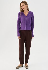 Cardigan en maille violette à col en V avec fermeture à boutons et motif texturé, associé à un pantalon marron taillé, avec des accents de poches décoratives.