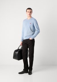 Helles blaues Strickpullover, schwarze Hose und schwarze Ledertasche. Mit Rundhalsausschnitt, gerippten Bündchen und einem kleinen roten Logo auf der Brust.
