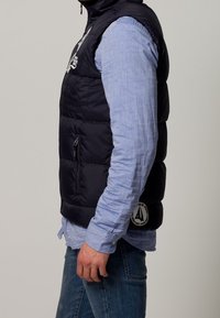 Marinblå pufferväst med quiltat mönster, hög krage och blixtlåsfickor. Har en logotyp på nedre högra sidan och är lager-på-lager över en ljusblå skjorta.