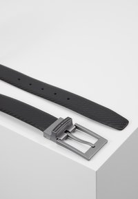 Ceinture synthétique noire avec une finition texturée, dotée d'une boucle en métal gris mat et de plusieurs trous d'ajustement le long de la sangle.