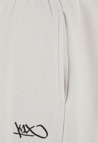 Lys grå joggere med en blød tekstur, der har sidelommer og et sort broderet logo nær kanten.