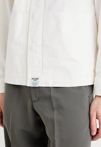 Camică albă cu nasturi, din bumbac, cu o textură netedă, având o etichetă la tiv și pantaloni gri croiți dedesubt.