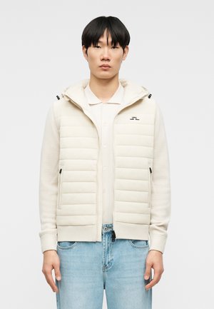 Fiatal férfi krémszínű steppelt, kapucnis cipzáras dzsekit visel, világos színű galléros inggel és kék farmerrel, egyszerű háttér előtt áll.