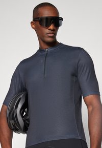 Maillot de cyclisme à manches courtes bleu marine avec fermeture éclair devant, fabriqué dans un matériau léger, associé à un casque noir et de grandes lunettes de soleil.