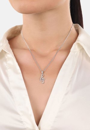 Silberne Halskette mit einem Design in Form eines Notenschlüssels, verziert mit kleinen, klaren Steinen, getragen über einem hellen Satin-Hemd.