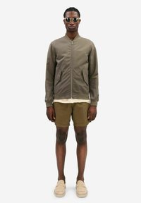 Olive green bomber jacket with a zip, two side pockets, paired with light beige T-shirt, brown shorts, and beige slip-on shoes. Sunglasses present.