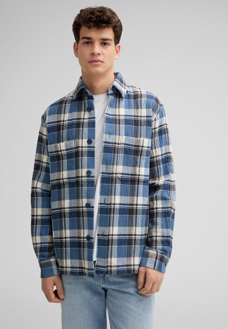 Flanellen overhemd in blauw, zwart en wit ruitpatroon met een gespreide kraag, knoopsluiting en borstzak, gecombineerd met een wit T-shirt eronder.