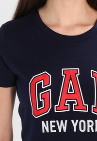 Námořnická modrá bavlněná tričko se silnými červenými a bílými nápisy "GAP NEW YORK", kulatým výstřihem a krátkými rukávy.