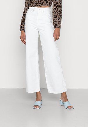 Kobieta ubrana w białe, szerokie spodnie z wysokim stanem, brązową, długorękawową bluzkę w kwiaty oraz niebieskie sandały na blokowym obcasie, stojąca na szarej podłodze.