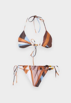 Žena v pestrofarebne pruhovanom bikinách a čiernom šatke na hlave, stojaca bosá na jednoduchom svetlom pozadí.